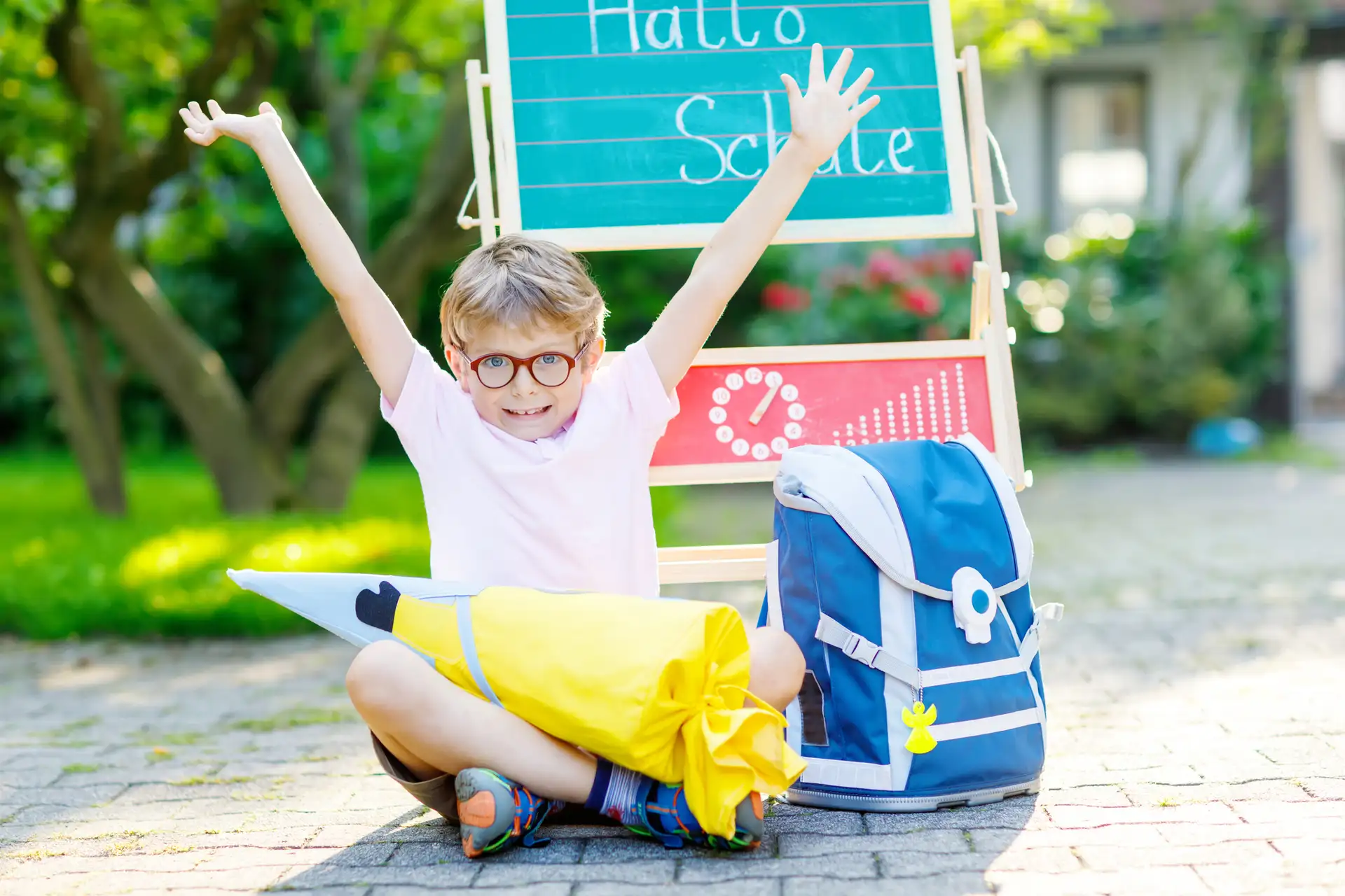 Happy little kid boy with glasses sitting by desk and backpack or satchel. Schoolkid with traditional German school bag called Schultuete on his first day to school. Hello school in German language