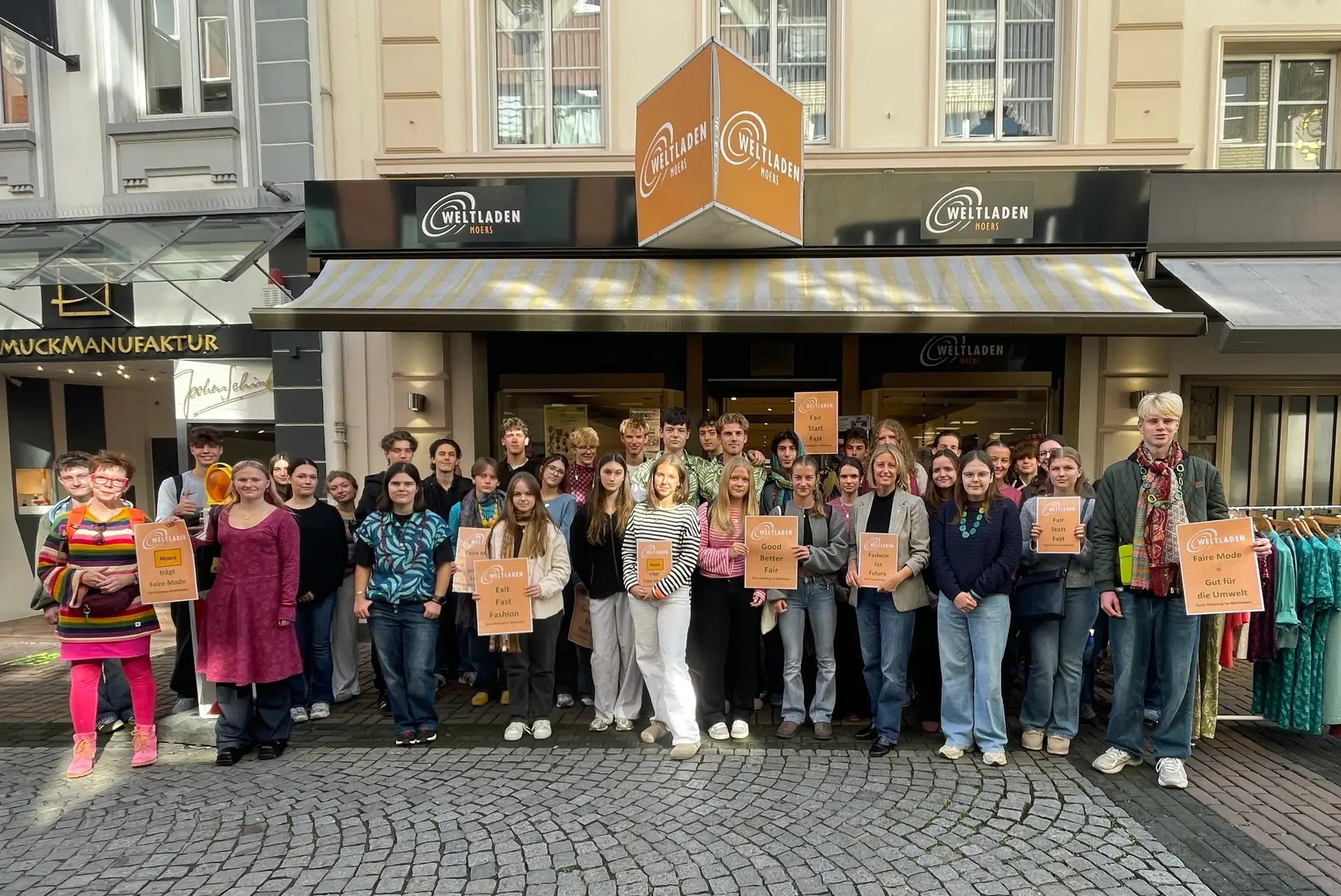 Die Bürgermeisterin wiegt sich im Weltladen auf einer Waage; im Hintergrund befinden sich Verkaufsregale mit fair gehandelten Produkten.