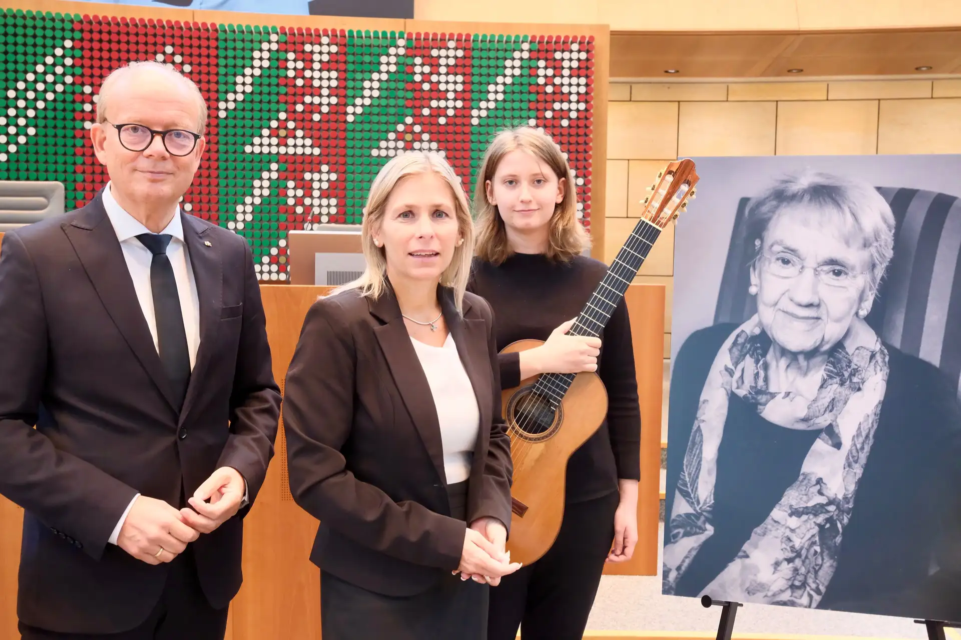 Landtagspräsident André Kuper, Bürgermeisterin Sonja Wienecke und Gitarristin Annika Schlitt stehen nebeneinander vor einem gemeinsamen Hintergrund und gedenken Ingeborg Friebe.