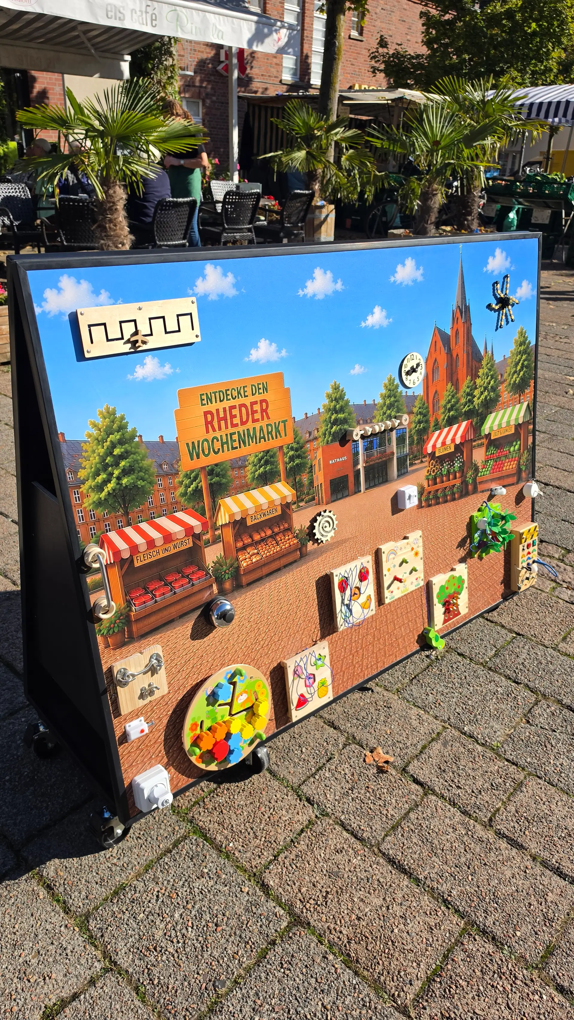 Activityboard mit einem gemalten Rheder Wochenmarkt und dem Rathaus.