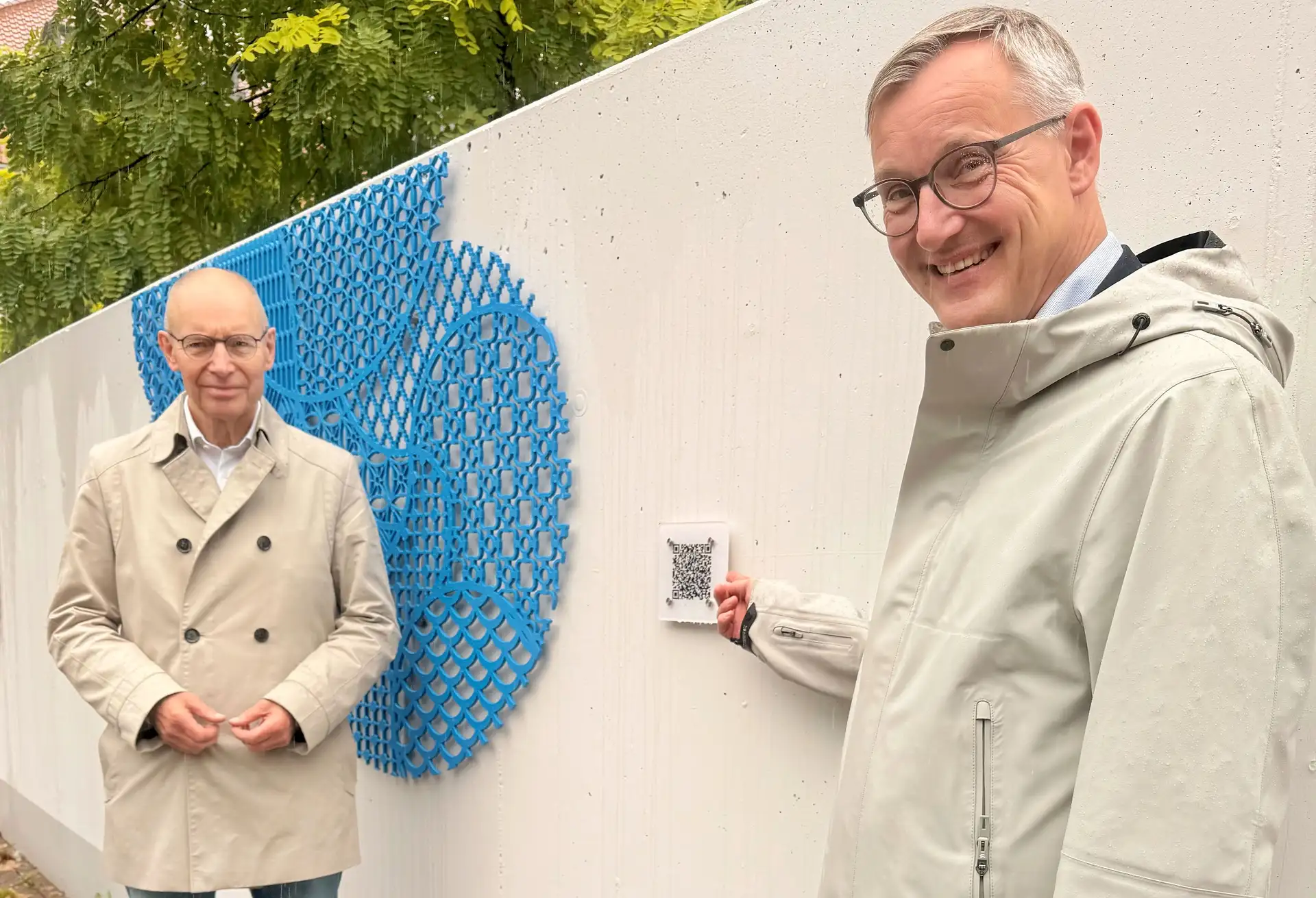Zwei Personen stehen nebeneinander und halten ein Pressefoto vor einer Installation mit QR-Codes und farbigen Wolken-Elementen.