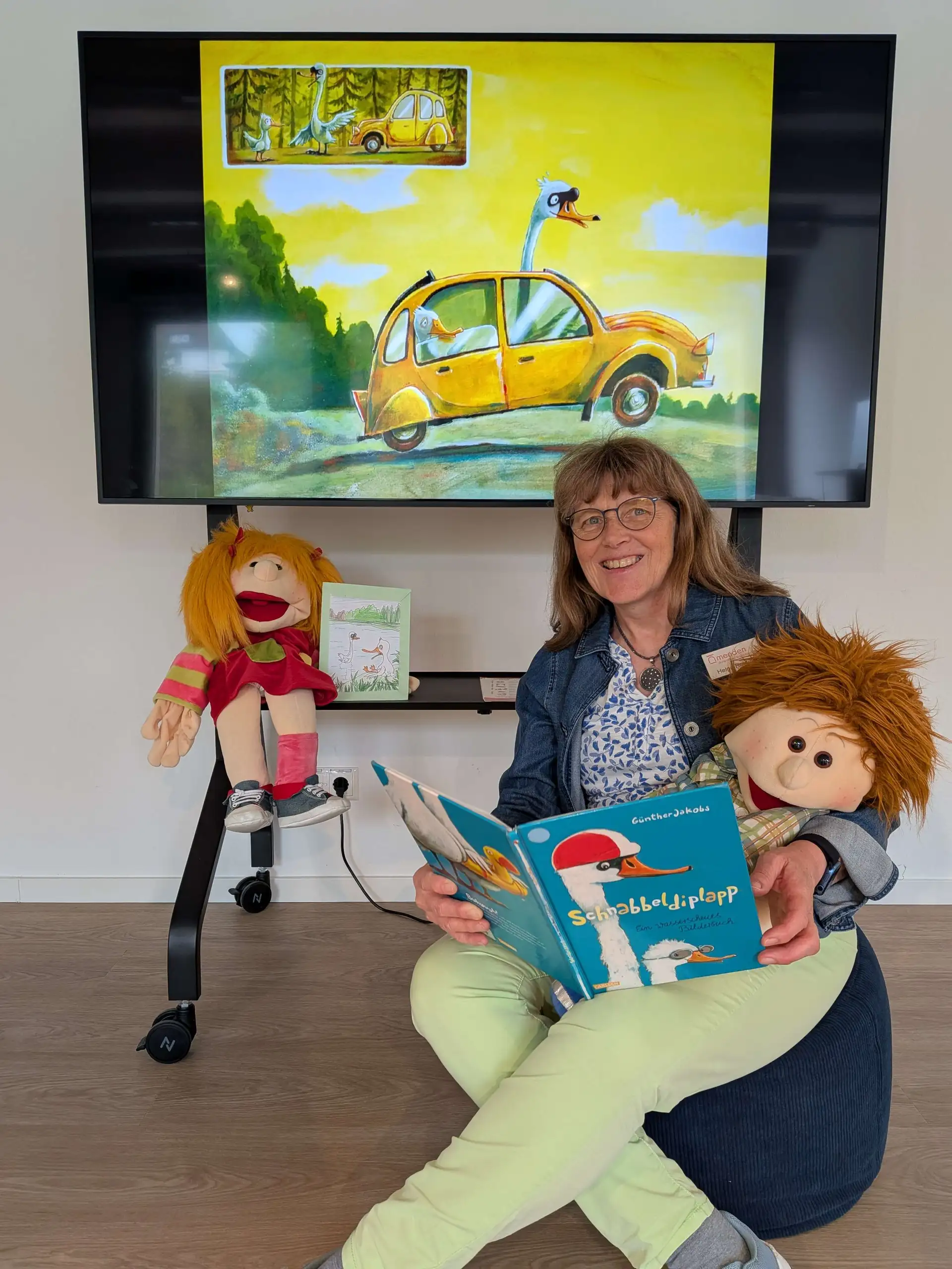 Helga Rebling hält ein aufgeschlagenes Bilderbuch. Neben ihr sind zwei Handpuppen und hinter ihr ein Bildschirm mit einer Illustration zu sehen.