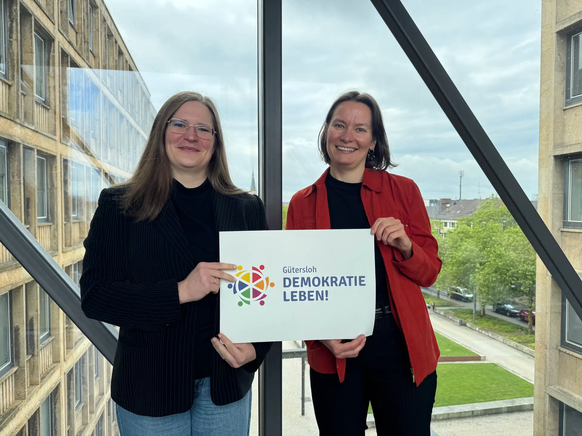 Zwei Personen stehen nebeneinander bei einer Veranstaltung und laden zur Demokratiekonferenz „Demokratie und Arbeitswelt“ ein.