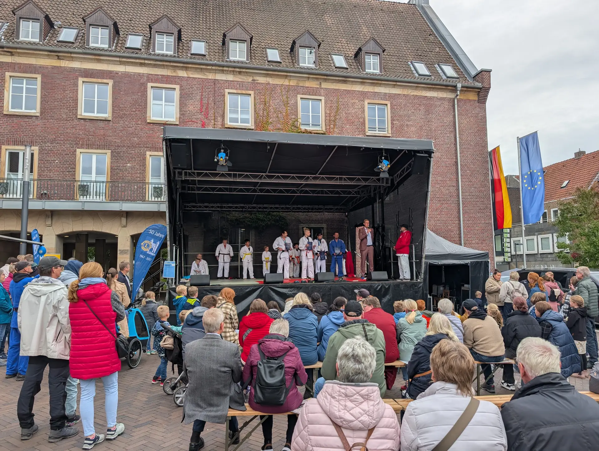 Menschen in Sportanzügen stehen auf einer Bühne vor dem Rathaus in Dülmen; davor sieht Publikum zu.